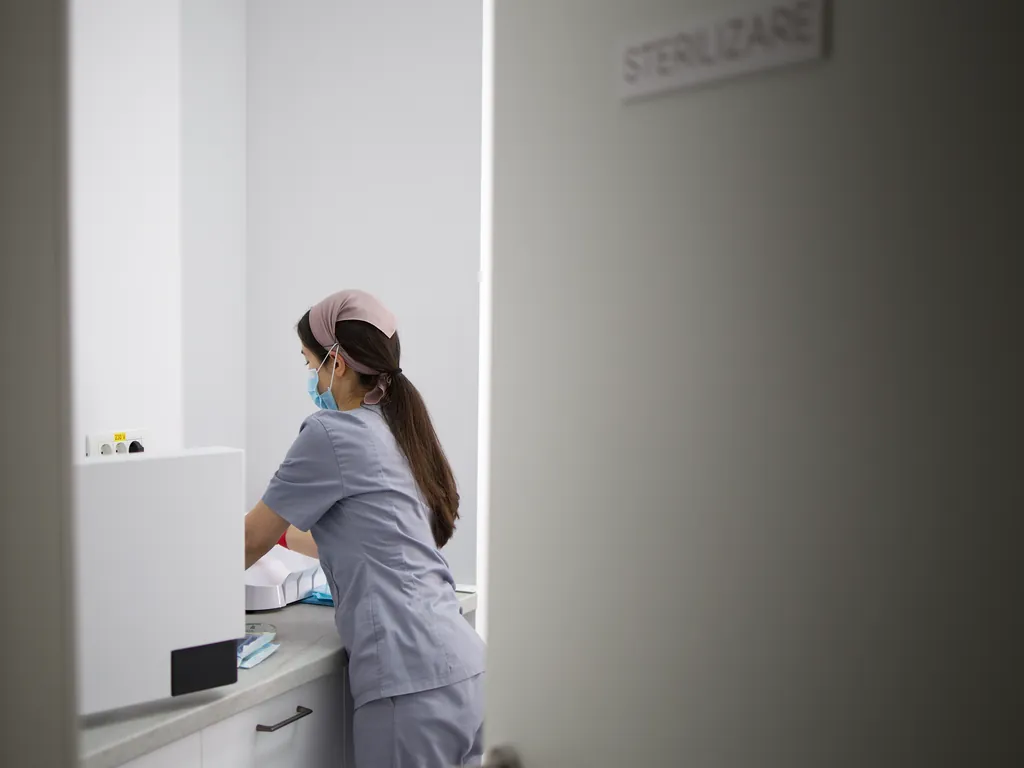 Sala de sterilizare văzută prin ușa întredeschisă, cu o colegă la aparatură