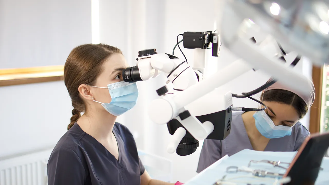 Medic stomatolog lucrând la microscopul dentar, cu o colegă alături